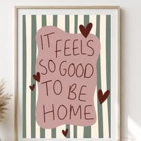 Hand-lettered print reading It feels so good to be home on dusky pink with sage stripes and hearts